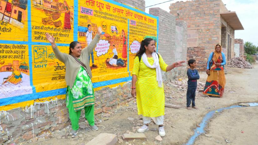 Three women and a child stand in front of a mural created by the Mahila Housing Trust in Jodhpur, India. The mural is part of a larger initiative on mitigating the impacts of extreme heat on vulnerable communities in Jodhpur through early warning systems and raising awareness.
